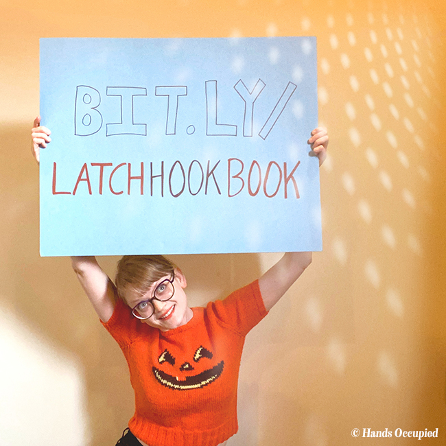 Heidi Gustad holds up a sign reading bit.ly/latchhookbook which is the URL find her first book