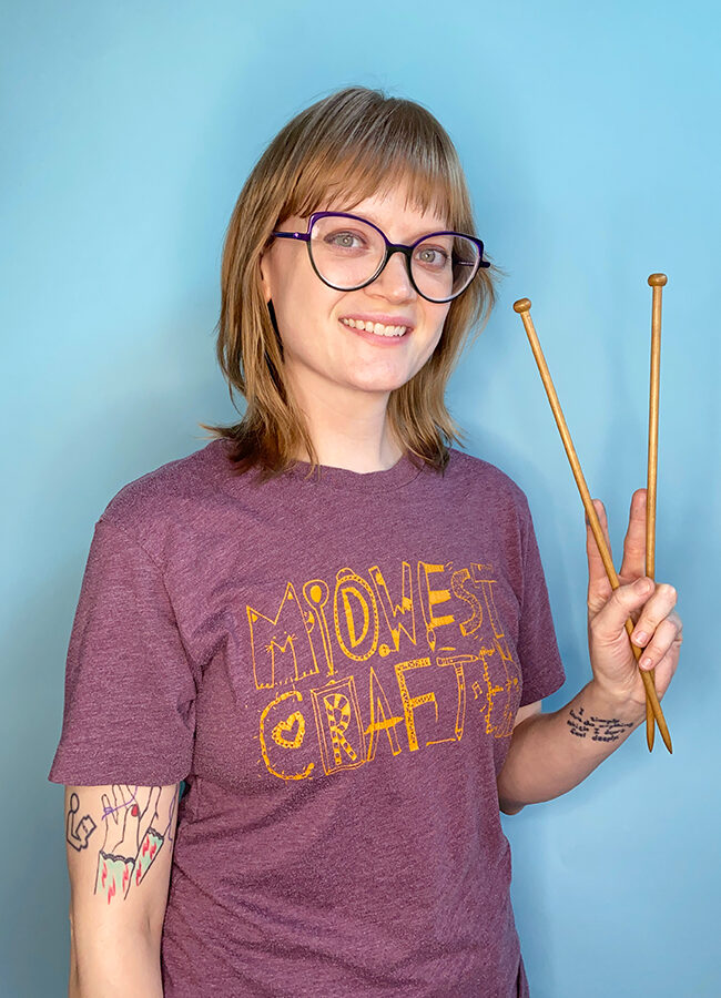 Heidi Gustad from Hands Occupied holds up a pair of wooden knitting needles. She's wearing a t-shirt that reads "Midwest Crafter."