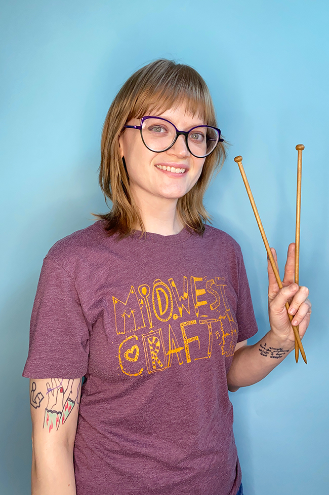 Heidi Gustad from Hands Occupied holds up a pair of wooden knitting needles. She's wearing a t-shirt that reads "Midwest Crafter." 
