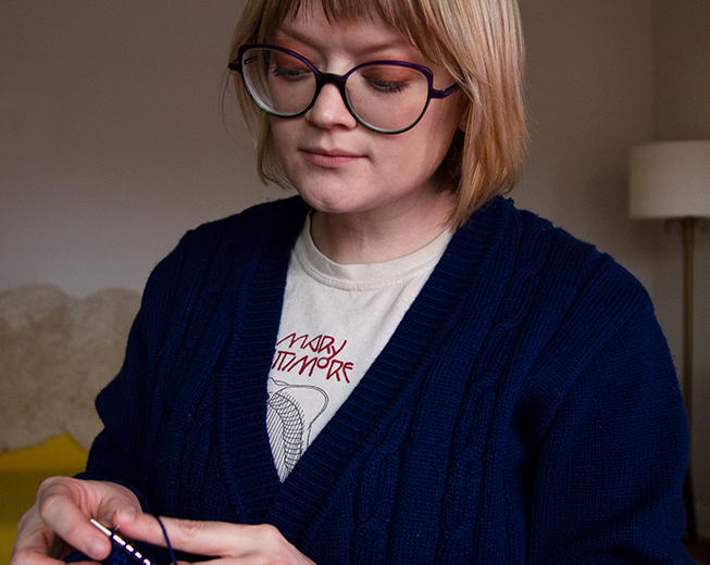 A blond woman with glasses and cardigan knits while looking down at her project.
